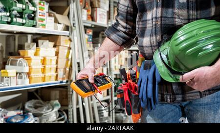 L'elettricista tiene a portata di mano il tester per multimetro, l'elmetto con occhiali di protezione. Industria edile, impianto elettrico. Foto Stock