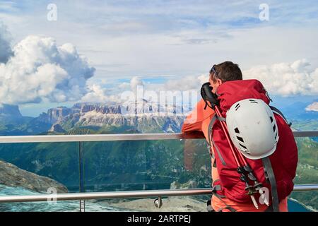 Uomo turistico con bastoni da trekking e casco da arrampicata sul suo zaino rivolto verso il Gruppo Sella in montagna Dolomiti. Vista dalla Terrazza Marmolada o Foto Stock