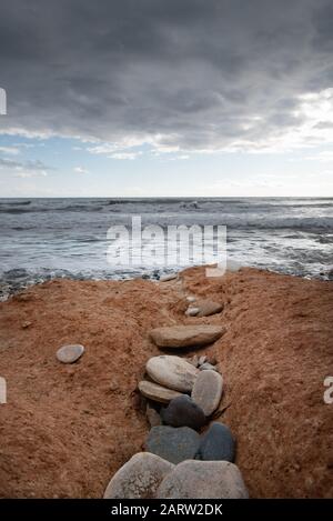 Fila di ciottoli su una spiaggia ondulata rossa e tempestosa cielo nuvoloso Foto Stock