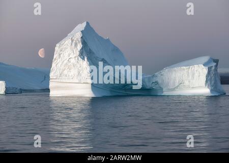 Enorme iceberg galleggiante in un fiordo al tramonto con la luna sulla sinistra. Scoresby Sund, Kangertitivaq, Groenlandia, Danimarca Foto Stock