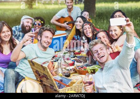 Gruppo di amici diversi che scattano foto selfie mentre fanno picnic nel parco - Giovani Felici che hanno con la nuova tecnologia del telefono mobile e mangiare Foto Stock