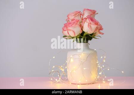 Vaso con rose fresche e ghirlanda luminosa sul tavolo Foto Stock