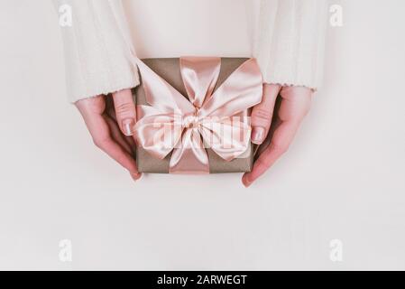 Le mani della donna tengono la scatola del regalo avvolta con carta artigianale e nastro rosa su sfondo rustico pastello. Foto Stock