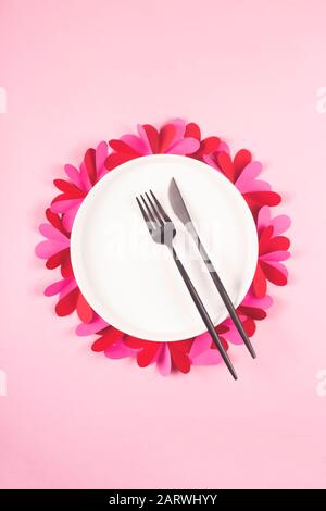 Piatto bianco rotondo con una forcella nera e un coltello su un tavolo rosa con cuori di carta. Concetto di San Valentino e tavolo posato. Foto Stock