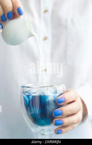 Matcha blu bevanda fredda con ghiaccio in un bicchiere di vetro nelle mani di una donna con manicure blu. Colore dell'anno 2020 trend Foto Stock