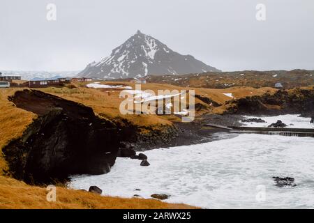 Fiume circondato da colline coperte di verde e neve in Un villaggio in Islanda Foto Stock