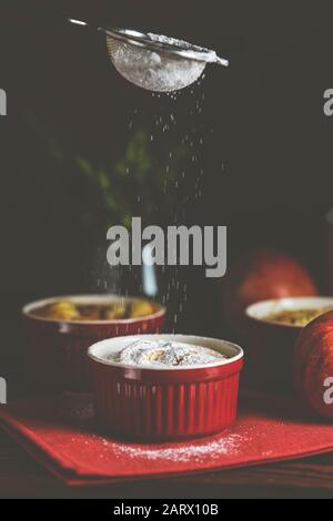 Setacciare lo zucchero a velo su torta di mele in stampi di cottura in ceramica ramekin su tavola di legno scuro. Primo piano, profondità poco profonda del campo. Foto Stock