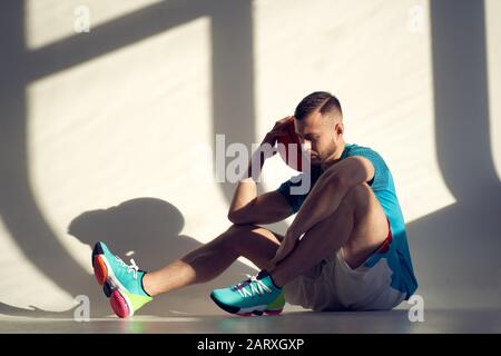 Giovane atletico, giocatore di pallacanestro che tiene la palla sulla spalla e seduto vicino al muro con le ombre dalla finestra Foto Stock