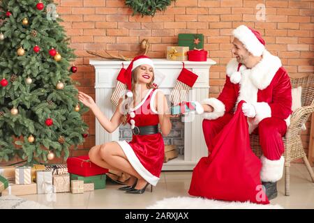 Giovane coppia vestita come Babbo Natale mettendo regali sotto abete in camera decorata per Natale Foto Stock