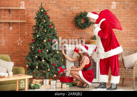Giovane coppia vestita come Babbo Natale mettendo regali sotto abete in camera decorata per Natale Foto Stock