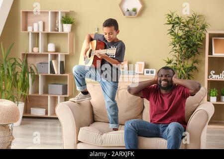 Uomo afro-americano che copre le orecchie mentre suo figlio piccolo sta suonando la chitarra a casa Foto Stock