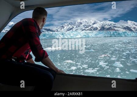 Il giovane uomo sta guardando su un bel ghiacciaio Hubbard. È su una nave da crociera in Alaska. Il maschio indossa una camicia lumberjack e guarda l'iceberg. Foto Stock