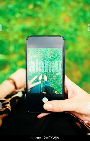 prospettiva personale pov di donna che scatta la fotografia con lo smartphone del telefono mobile con la mano e le gambe nella cornice Foto Stock