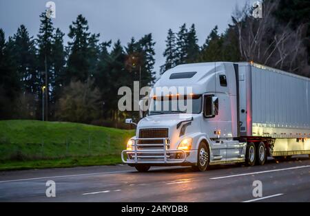 Grande carro industriale diesel bianco semi-camion con griglia di protezione che trasporta il carico commerciale in furgone secco semi-rimorchio che corre sulla strada bagnata serale con Foto Stock