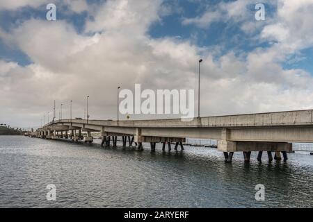 Oahu, Hawaii, Stati Uniti. - 10 Gennaio 2020: Pearl Harbor. Il ponte chiatta dell'isola Ford fruma la linea di cemento beige tra l'acqua grigia e il paesaggio blu. Foto Stock