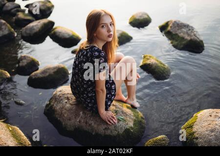Donna che indossa un abito nero seduto sulla roccia in mare Foto Stock