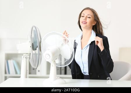 Giovane donna che usa ventilatori elettrici durante l'ondata di caldo in ufficio Foto Stock