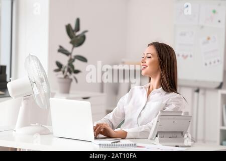 Giovane donna che usa un ventilatore elettrico durante l'ondata di caldo in ufficio Foto Stock