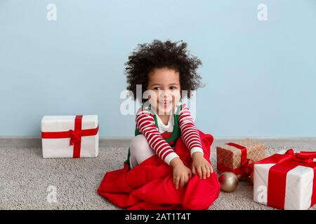 Bambina in costume da elfo, con borsa di Babbo Natale e regali di Natale seduti vicino alle pareti colorate Foto Stock