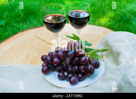 Due bicchieri di vino rosso e un grappolo d'uva su un tavolo di legno su uno sfondo di erba verde. Foto Stock