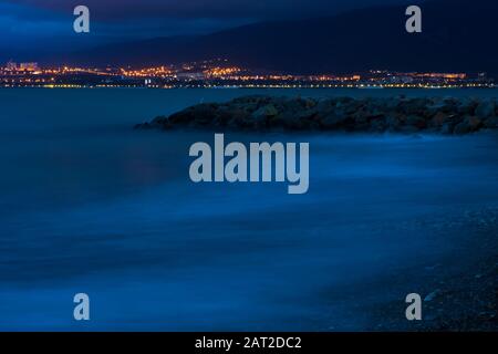 Surf sul mare in una lunga esposizione in serata. Le onde rotolano sopra la spiaggia di ciottoli e le rocce. Sullo sfondo, montagne e luci della città. Esposizione lunga. Foto Stock