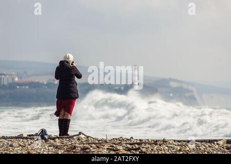 una ragazza fotografa grandi onde tempeste sullo sfondo di un faro e rocce. La ragazza è vestita con una giacca nera, cappello bianco lavorato a maglia, Borgogna l Foto Stock