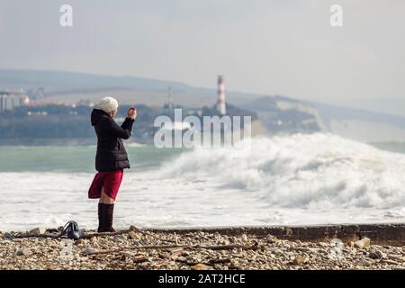 una ragazza fotografa grandi onde tempeste sullo sfondo di un faro e rocce. La ragazza è vestita con una giacca nera, cappello bianco lavorato a maglia, Borgogna l Foto Stock