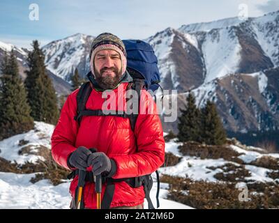 Turista barbuto in inverno. Un uomo con uno zaino e una calda giacca cremisi Foto Stock