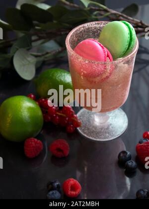 Wineglass rosa con macaron multicolore, lime e bacche fresche diverse sparse su una superficie specchio Foto Stock