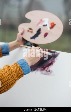 Primo piano foto di giovane artista femminile che lavora sulla pittura all'aperto. Contiene vernici ad olio, pennelli per artisti, tele e palette. Sta mescolando i colori sopra Foto Stock