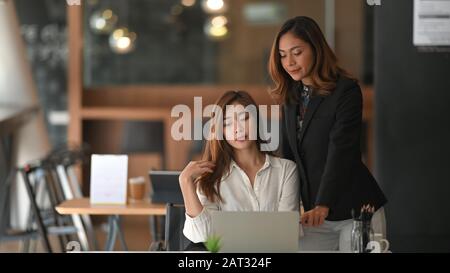 Donna sicura di affari in abito nero in piedi e consigliando a bella camicia bianca dipendente che seduta / posa sul moderno sfondo dell'ufficio. Foto Stock