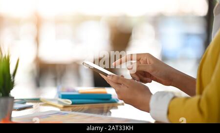 Vista laterale di un allegro designer che utilizza uno smartphone nero a portata di mano e punta il dito su di esso mentre si siede alla scrivania in legno. Foto Stock