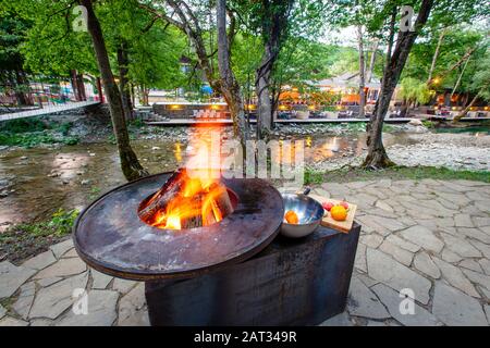 Barbecue con un fuoco acceso sulla riva del fiume di montagna la sera. Accanto alla griglia verdure preparate per friggere: Peperoni, pomodori, mu Foto Stock