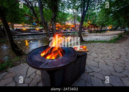 Barbecue con un fuoco acceso sulla riva del fiume di montagna la sera. Accanto alla griglia verdure preparate per friggere: Peperoni, pomodori, mu Foto Stock