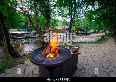 Barbecue con un fuoco acceso sulla riva del fiume di montagna la sera. Accanto alla griglia verdure preparate per friggere: Peperoni, pomodori, mu Foto Stock