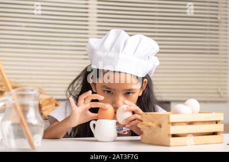 Una bambina asiatica che indossa un cappello bianco dello chef sta fissando le uova sul piccolo brocca e mette su un tavolo da cucina bianco in cucina. Foto Stock