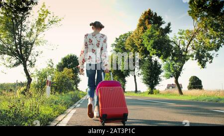 Donna allegra con valigia che cammina su strada. Da sotto scatto di bella giovane femmina sorridente e tirando valigia rossa mentre si cammina su strada asfaltata durante il viaggio attraverso la campagna. Foto Stock