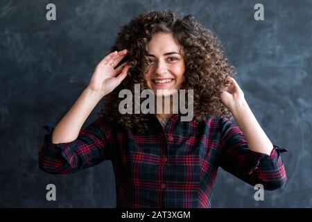 Primo piano scatto studio di una ragazza bruna gioiosa curly contro la parete grigia, toccando i capelli.- immagine Foto Stock