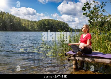 Giovane donna incinta vestita sportivo fare esercizio di yoga seduto in posizione lotus su un molo di legno in un lago di foresta meditando. Fitness e guarire Foto Stock