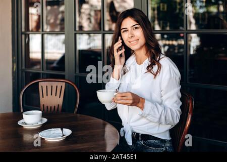 La ragazza sorridente in camicia bianca tiene la tazza e parla nel caffè Foto Stock