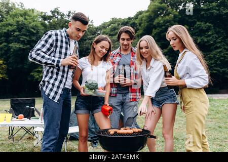 Una compagnia di amici con un drink e cucina sul barbecue nel campeggio . - Immagine Foto Stock