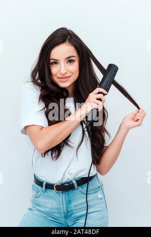 Giovane donna sorridente che lisciava i capelli con una piastra sullo sfondo bianco Foto Stock