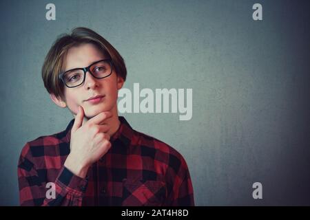 Ragazzo adolescente sicuro di pensare, tiene una mano sotto chinlooking via isolato su sfondo grigio parete con spazio copia. Sognatore ragazzo studente adolescente Foto Stock