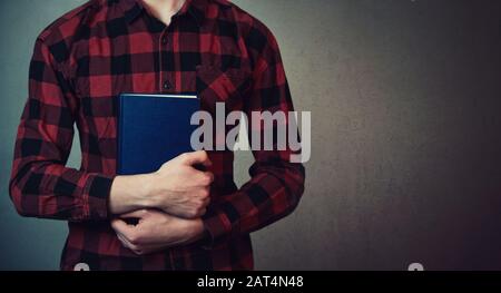 Primo piano di ragazzo adolescente che tiene un libro blu vicino al corpo. Concetto di istruzione come studente abbracciando un libro di testo, amorevole conoscenza isolato sulla parete grigia bac Foto Stock