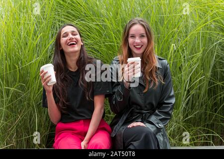 Due donne di diverse etnie che tengono tazze usa e getta all'aperto e sorridono alla macchina fotografica. Foto Stock