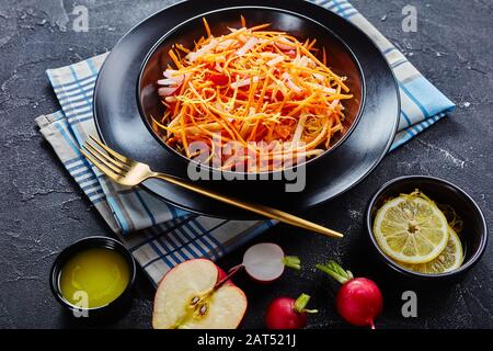 Primo piano di ravanello di carote Fresche e insalata di mele cosparse di semi di quinoa e scorza di limone in una ciotola nera su un tavolo di legno con condimento fatto in casa Foto Stock