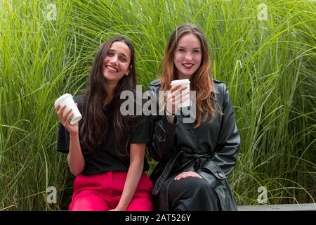 Ritratto di due diverse giovani donne con tazze usa e getta guardando la fotocamera con sorrisi. Erba sfondo. Foto Stock