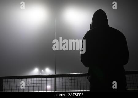 Una misteriosa figura con cappuccio, in piedi con la schiena alla fotocamera su un ponte, guardando fuori le luci di strada. Durante una notte di nebbia. Foto Stock