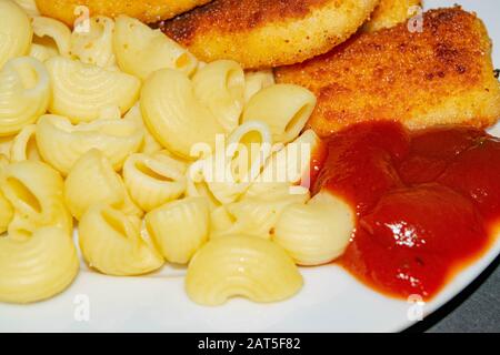 Pepite di pollo fritte e pasta bollita con ketchup su un piatto. Primo piano Foto Stock