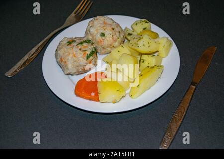 Due polpette di carne con patate bollite e carote bollite su un piatto bianco con un coltello e forchetta su uno sfondo scuro. Primo piano Foto Stock